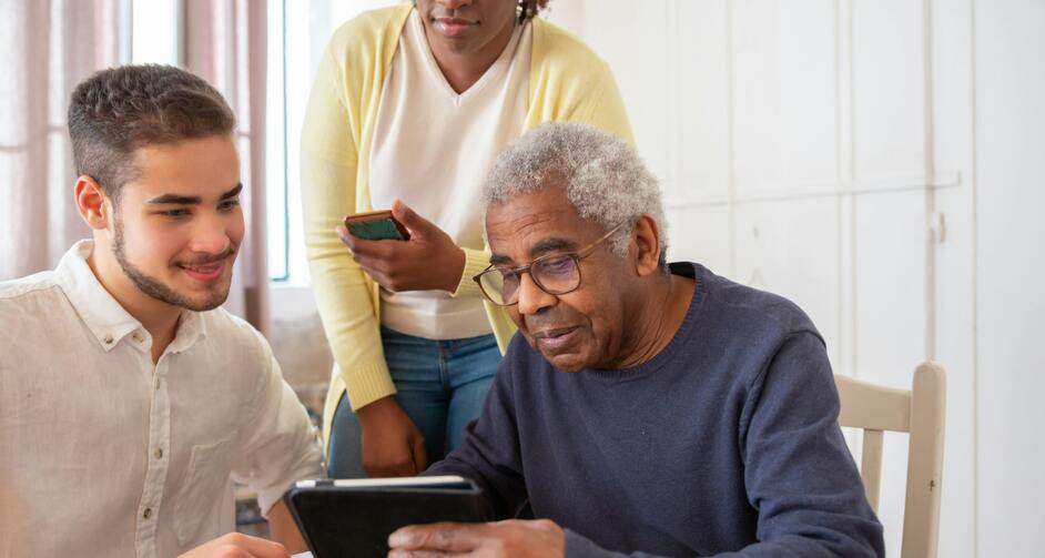 Older man and younger man seated at table with younger woman standing behind, all looking at a tablet the older man is holding