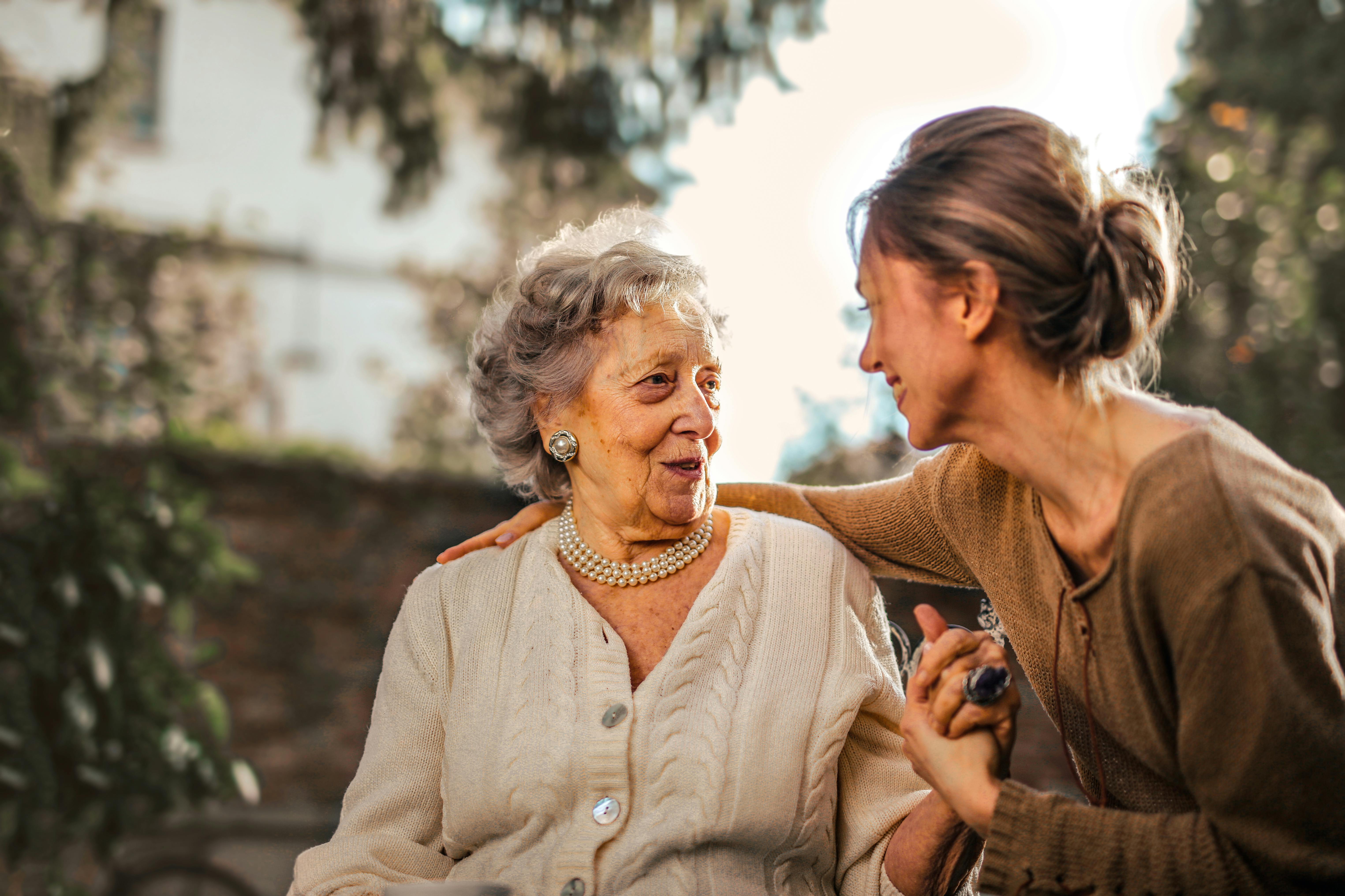 Older woman seated outdoors holding hands and talking to younger woman with arm around older woman's shoulders