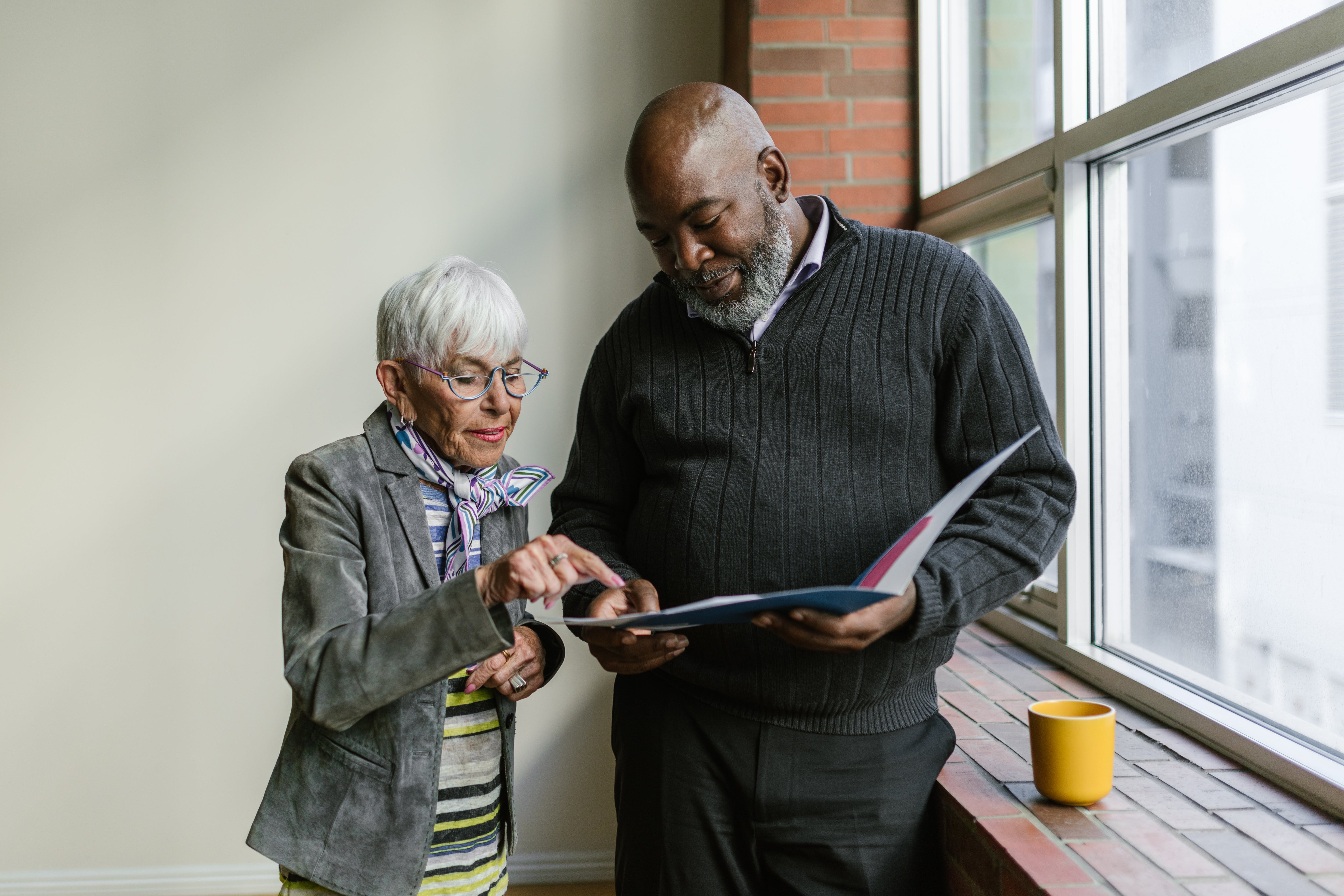 Older man and older woman standing next to large window while reading an open folder, woman is pointing to something in the folder