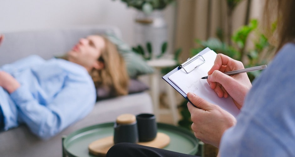 Woman holding clipboard writing while listening to man lying down on couch